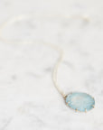 A delicate silver chain necklace with a round, light blue stone pendant set in a gold-toned bezel, resting on a white marble surface.