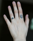 A close-up of a person's hand showing a silver ring with a dark oval gemstone on the ring finger, alongside a small geometric tattoo on the middle finger.