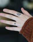 A close-up of a person's hand wearing a delicate gold ring with a small round blue gemstone, set against a dark blurred background and a brown knit sweater cuff.