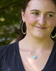 A woman wearing a silver necklace with a light blue stone pendant, gold hoop earrings, and a nose ring, photographed outdoors against a blurred green background.