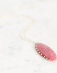 A delicate gold chain necklace with an oval pink stone pendant, resting on a light marble surface.