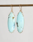 A pair of gold drop earrings with oval light blue turquoise-like stones, hanging from a horizontal gold rod against a plain light gray background.