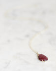 A delicate gold chain necklace with a small, teardrop-shaped deep red gemstone pendant, resting on a light marble surface.