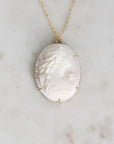 A gold chain necklace with an oval carved cameo pendant, displayed on a light marble surface. The pendant features a profile of a woman in cream and white with gold prong settings.
