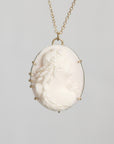 A gold chain necklace featuring an oval white carved cameo pendant with a classical profile design, set in a gold bezel with small decorative side prongs, shown against a neutral light gray background.
