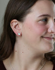 A close-up of a woman's ear wearing a small round silver stud earring with a clear gemstone, along with other ear piercings. The background is a soft, neutral gray.