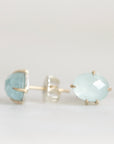 A pair of oval-cut light blue gemstone stud earrings in gold settings, with one earring shown from the front and the other from the side/back, on a plain light background.