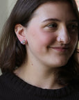 A close-up portrait of a woman with brown hair wearing a small silver hoop earring with a clear gemstone, shown from the chest up against a softly blurred indoor background.