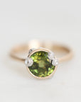 A close-up shot of a ring featuring a round green gemstone in a gold bezel setting, accented with three small clear stones on the sides, set against a plain light gray background.