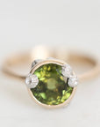 A close-up shot of a ring featuring a round green gemstone in a gold bezel setting, accented with three small clear stones on the sides, set against a plain light gray background.