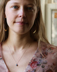 Woman wearing opal necklace with gold chain. She is wearing a floral top and large gold hoops. Indoor background.