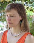 Woman wearing a pearl necklace and orange top with greenery in the background