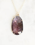 A teardrop-shaped purple gemstone pendant with gold prongs, hanging from a delicate gold chain on a white marble background.