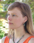 Woman with long blonde hair wearing an orange top and a necklace, standing outdoors with greenery in the background.