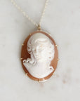 An oval pendant necklace featuring a carved white classical bust on a warm brown background, set in a gold-toned frame and hanging from a delicate chain on a light marble surface.