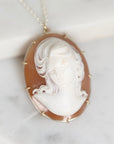 An oval pendant necklace featuring a carved white classical bust on a warm brown shell-like background, set in a gold-toned bezel with small gold spikes, hanging from a delicate chain on a light marble surface.