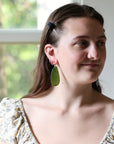 A woman wearing a large, organic-shaped olive green drop earring, shown in a close-up portrait against a softly blurred window background.