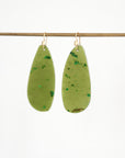 A pair of teardrop-shaped green resin earrings with tiny green inclusions and gold-tone hooks, hanging from a thin horizontal rod against a plain white background.