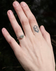 Hand with a ring and tattoo against a dark green leafy background