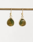 A pair of gold drop earrings with faceted green teardrop-shaped gemstones, hanging from a gold rod against a light gray background.