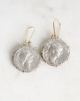 A pair of dangling earrings made from silver-toned ancient coin faces set in gold-toned metal hooks, displayed on a light marble surface.
