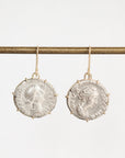 A pair of dangling earrings shaped like aged silver coins with gold-toned hooks, hanging from a horizontal gold rod against a plain light background.