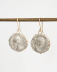A pair of dangling earrings with round silver-toned coin-style medallions in prong settings and gold-toned hooks, hanging from a horizontal rod against a plain light background.