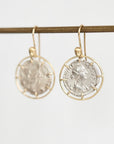 A pair of gold-tone dangle earrings with circular wire frames holding antique silver coins, hanging from a gold rod against a light gray background.