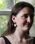 A woman with long brown hair is shown in profile, wearing a silver hoop earring with a white oval stone and a delicate silver necklace. She is wearing a black top, and the background is a softly blurred window with green foliage outside.