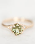 A close-up shot of a rose gold ring featuring a round-cut light green gemstone in a prong setting, set against a soft, out-of-focus white background.