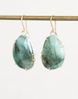 A pair of teardrop-shaped green stone drop earrings with gold hooks and prong settings, hanging from a gold rod against a light off-white background.