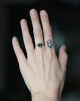 A close-up of a person's hand wearing a small gold ring with a green gemstone, with a geometric tattoo on the middle finger, against a dark blurred background.