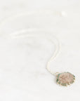 A delicate silver chain necklace with a small, scalloped circular pendant featuring a textured, frosted stone in muted green, pink, and beige tones, resting on a light marble surface.