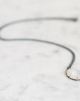 A delicate dark gray chain necklace with a small oval pendant featuring a milky white cabochon stone in a gold-toned bezel, resting on a white marble surface.