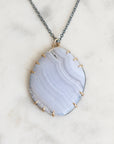 A delicate necklace featuring a light blue-gray agate stone pendant with gold prong settings and a dark gray cord, resting on a white marble surface.