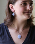 A woman wearing a matching jewelry set featuring a light blue-gray seashell pendant necklace and matching shell drop earrings on a silver chain.
