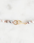 A close-up of a delicate jewelry piece featuring baroque pearls in silver, white, and gray tones, accented with rose gold wire and a gold hook-shaped charm, set against a light marble background.