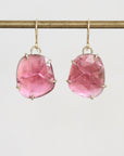 A pair of gold drop earrings with large, faceted pink gemstones in a prong setting, hanging from a gold rod against a light gray background.