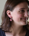 A woman with brown hair wearing a small pink dangling earring, shown in a close-up side profile against a soft neutral indoor background.