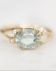 A yellow gold ring featuring a large oval-cut light blue gemstone in a prong setting, accented by two small round clear stones on the band, photographed on a soft white background.
