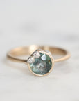 A gold ring featuring a round, faceted light blue-green gemstone with small barnacless on the upper left, set against a soft, light gray background.