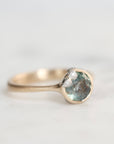 A close-up shot of a gold ring featuring a round light blue gemstone and a small cluster of barnacles on the band, set against a soft, light gray background.