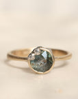 A silver ring featuring a round, faceted gray-green gemstone with small barnacles clustered along the top, set against a soft, light beige background.