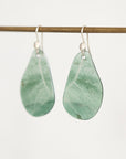 A pair of teardrop-shaped green stone pendants with silver fishhook ear wires, hanging from a thin horizontal wooden rod against a plain light background.