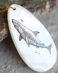 A white mother-of-pearl pendant necklace featuring a detailed black ink sketch of a shark, hanging from a gold chain and resting on a textured rock.