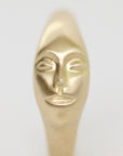 A close-up of a metallic gold object with a stylized human face sculpted on it, shown against a plain light gray background.