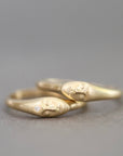 Two gold rings with sculpted faces resting on each other, one featuring a small clear gemstone, photographed on a neutral gray background.