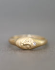 A gold ring featuring a stylized face design with a small clear gemstone on the side, photographed against a neutral gray background.