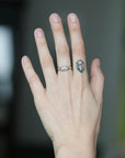 A close-up of a person's hand showing a silver ring with a small geometric cube design on the ring finger, set against a blurred dark background.