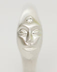 A close-up of a silver-toned sculptural face pendant or charm with closed eyes and a small clear gemstone set above the forehead, shown against a plain light gray background.